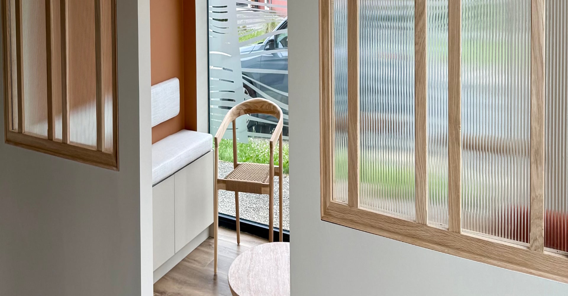 Intérieur moderne avec cloison en verre cannelé et bois clair. Vue sur banquette orange, meuble bas et chaise en bois près d'une fenêtre décorée.