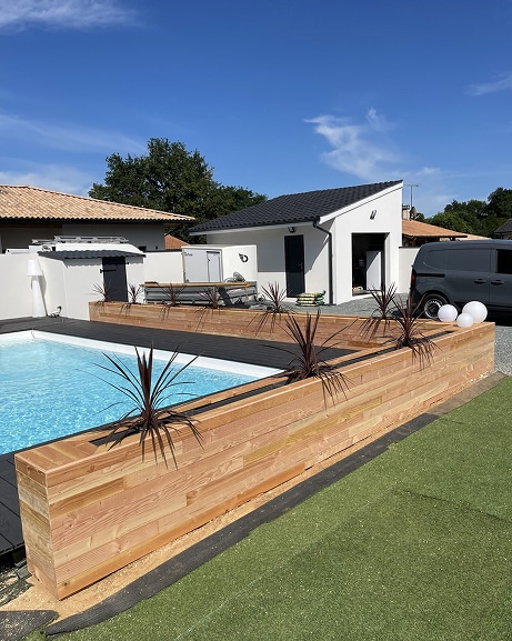 Piscine moderne avec terrasse en bois foncé et jardinière intégrée, gazon synthétique. En fond, pool house blanc et ciel bleu.
