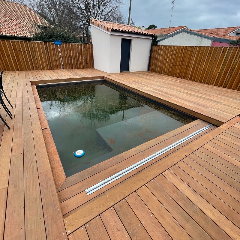 Piscine rectangulaire avec eau trouble, entourée d'une terrasse en bois neuve. Abri de jardin blanc et clôture bois.