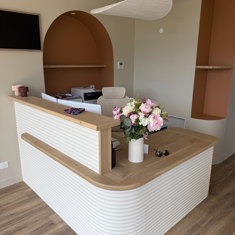 Réception moderne: bureau blanc nervuré, plateau bois clair, bouquet pivoines roses. Arches murales terracotta en fond.