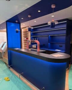 Intérieur de bar moderne avec comptoir bleu courbé, robinets de bière roses affichant 'VAPOTE', étagères bleues éclairées par LED et suspensions roses uniques.