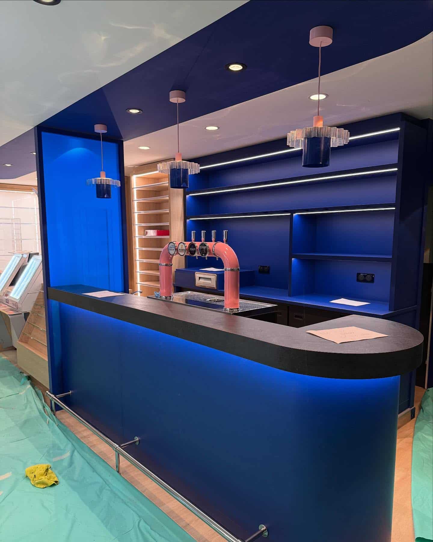 Intérieur de bar moderne avec comptoir bleu courbé, robinets de bière roses affichant 'VAPOTE', étagères bleues éclairées par LED et suspensions roses uniques.