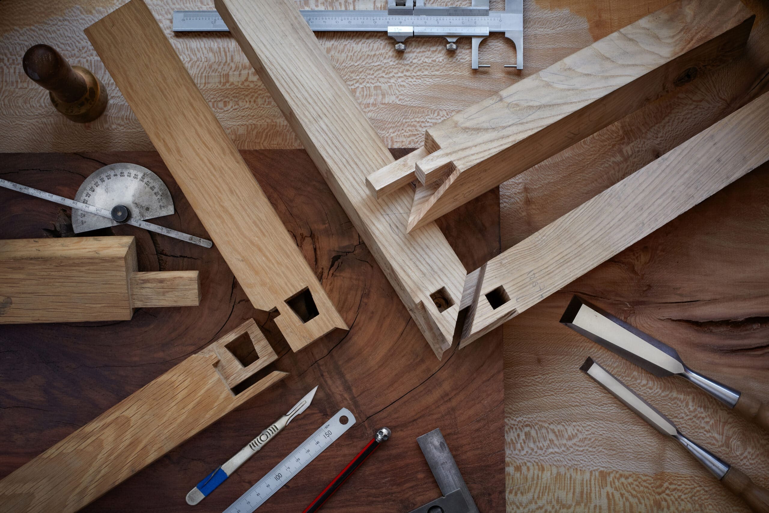 Outils et pièces de chêne clair préparées pour menuiserie fine sur un fond de bois foncé. Pied à coulisse, rapporteur, ciseaux, maillet. Assemblages bois à tenons.
