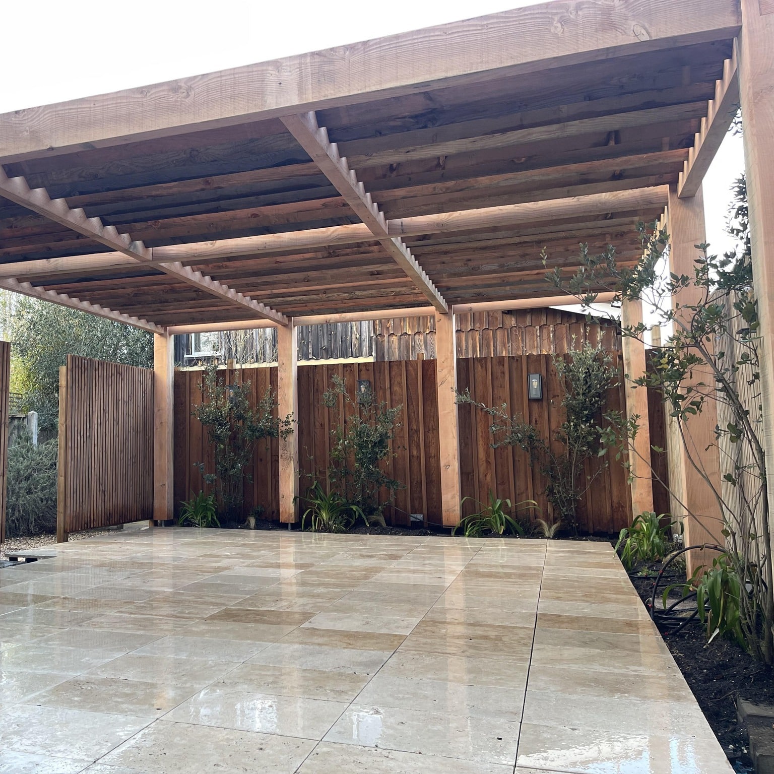 Grande terrasse carrelée beige brillante sous une pergola en bois clair et robuste, bordée d'une clôture et d'arbustes.