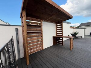 Terrasse moderne avec pergola en bois foncé, bar extérieur intégré et mur latté avec plantes. Decking gris et ciel bleu.
