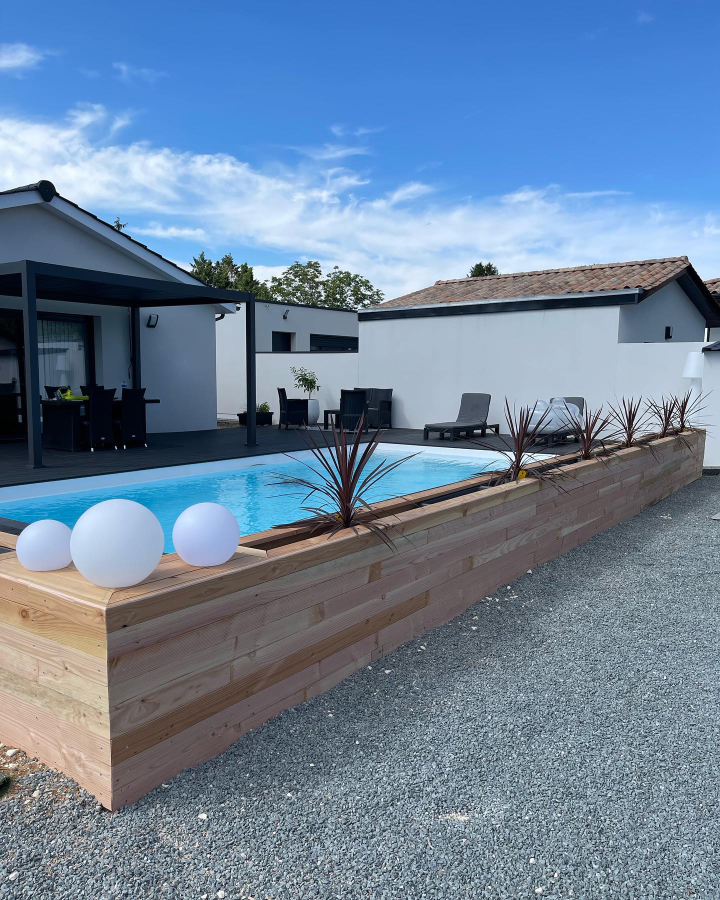 Piscine moderne avec margelle en bois et lumières sphériques dans un jardin paysager. Maison contemporaine et allée en gravier sous ciel bleu.