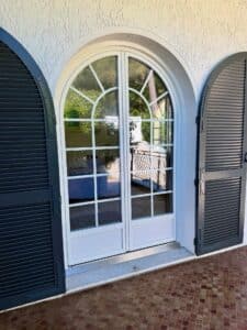 Élégante porte arquée blanche avec croisillons, flanquée de volets noirs à persiennes ouverts, sur un sol en carrelage brun texturé.