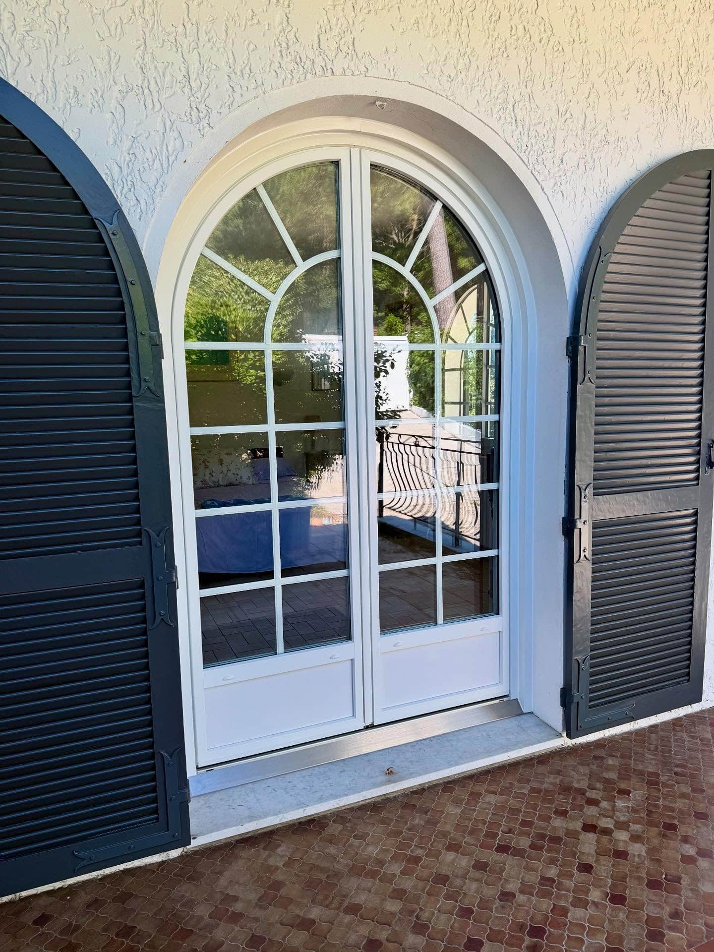 Élégante porte arquée blanche avec croisillons, flanquée de volets noirs à persiennes ouverts, sur un sol en carrelage brun texturé.
