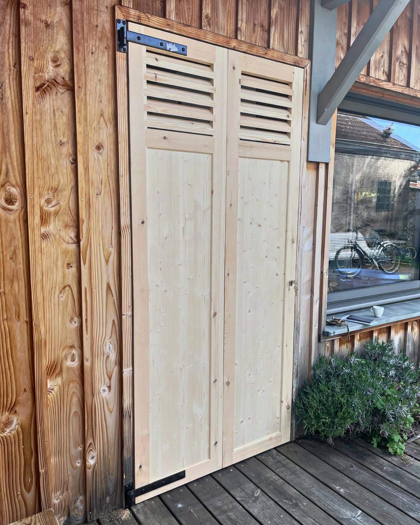 Porte double en bois clair à persiennes sur façade en bois rustique. Fenêtre à droite reflétant un vélo et une maison. Terrasse en bois, plantes.