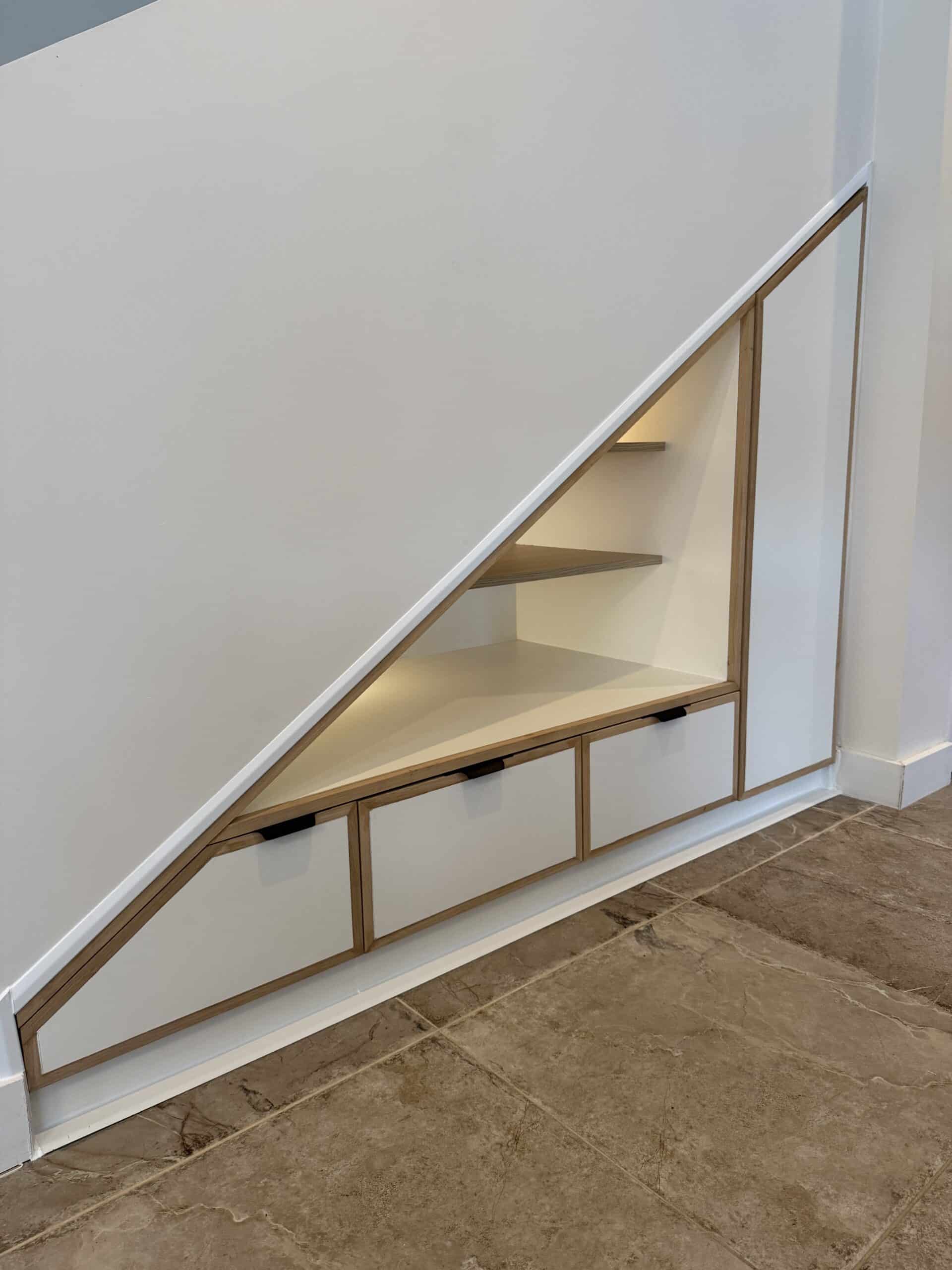 Rangement intégré moderne sous escalier avec trois tiroirs blancs, étagères en bois clair et éclairage LED.