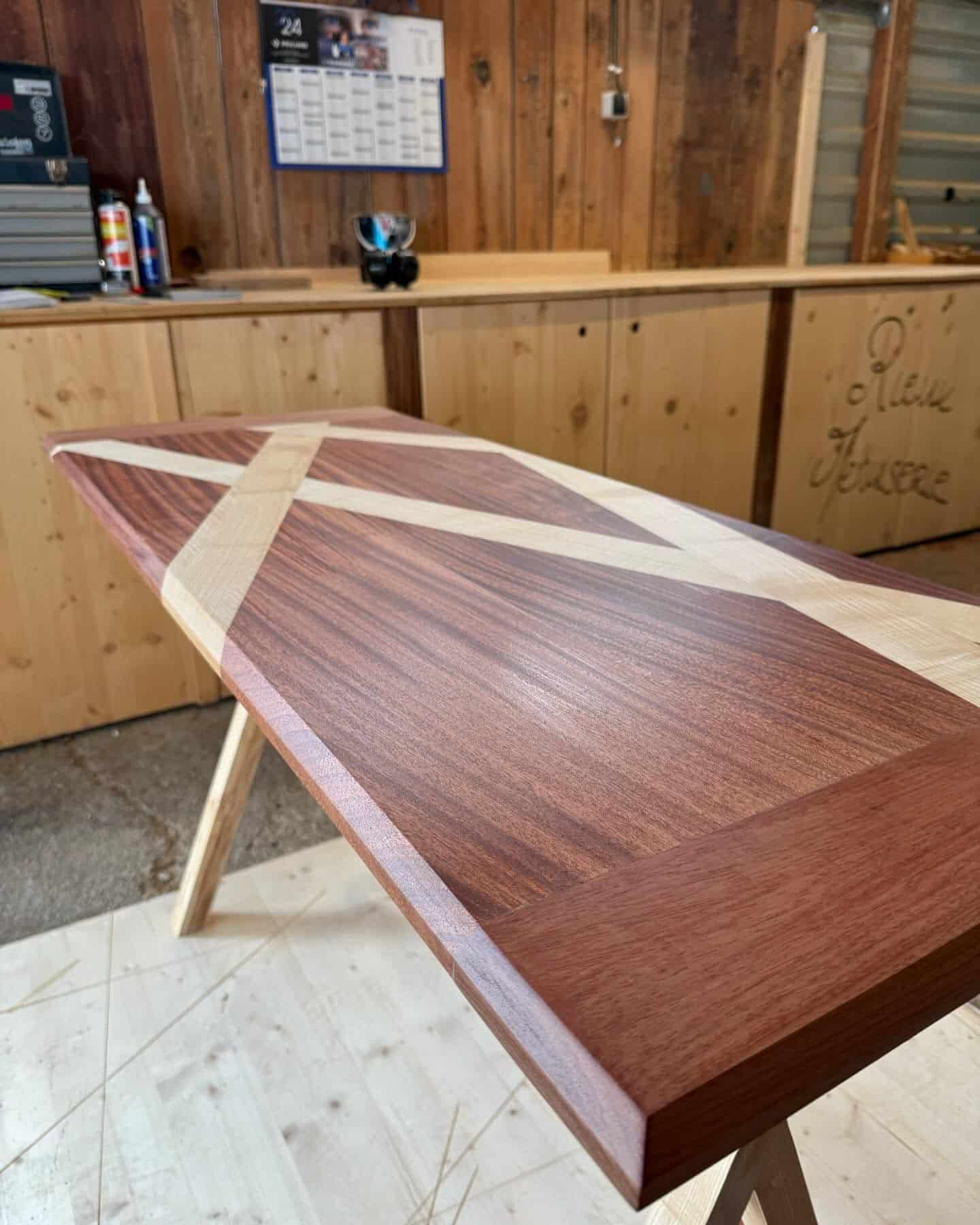 Table artisanale en bois foncé et clair, plateau marqueté avec motif en croix. Arrière-plan d'atelier avec text "Rêve Menuiserie".