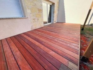 Terrasse en bois exotique rouge vif, fraîchement installée devant une maison moderne avec mur en pierre et volet roulant.