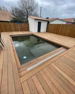 Nouvelle terrasse en bois clair autour d'une piscine rectangulaire avec rails de couverture et pool house. Jardin clôturé.