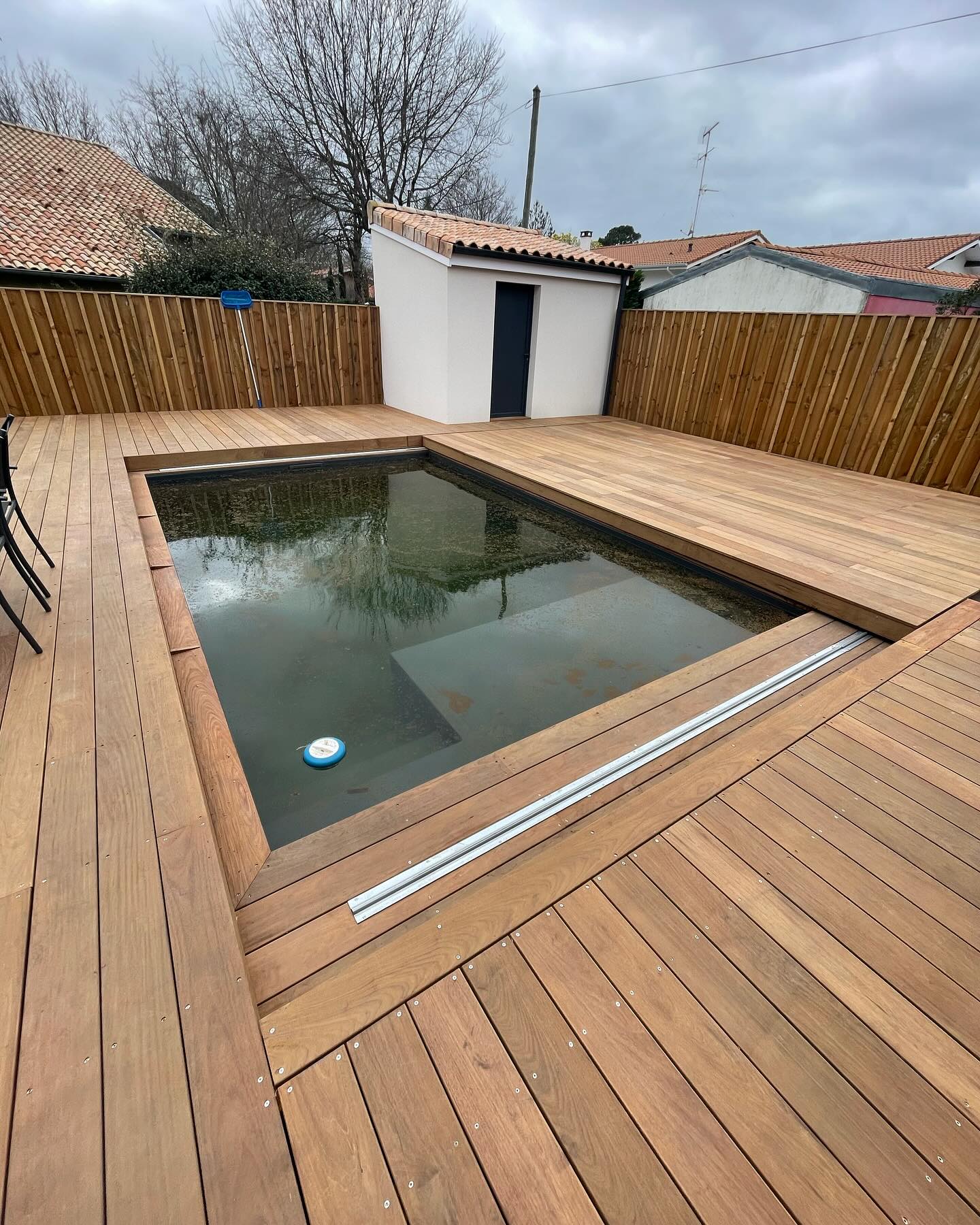 Nouvelle terrasse en bois clair autour d'une piscine rectangulaire avec rails de couverture et pool house. Jardin clôturé.