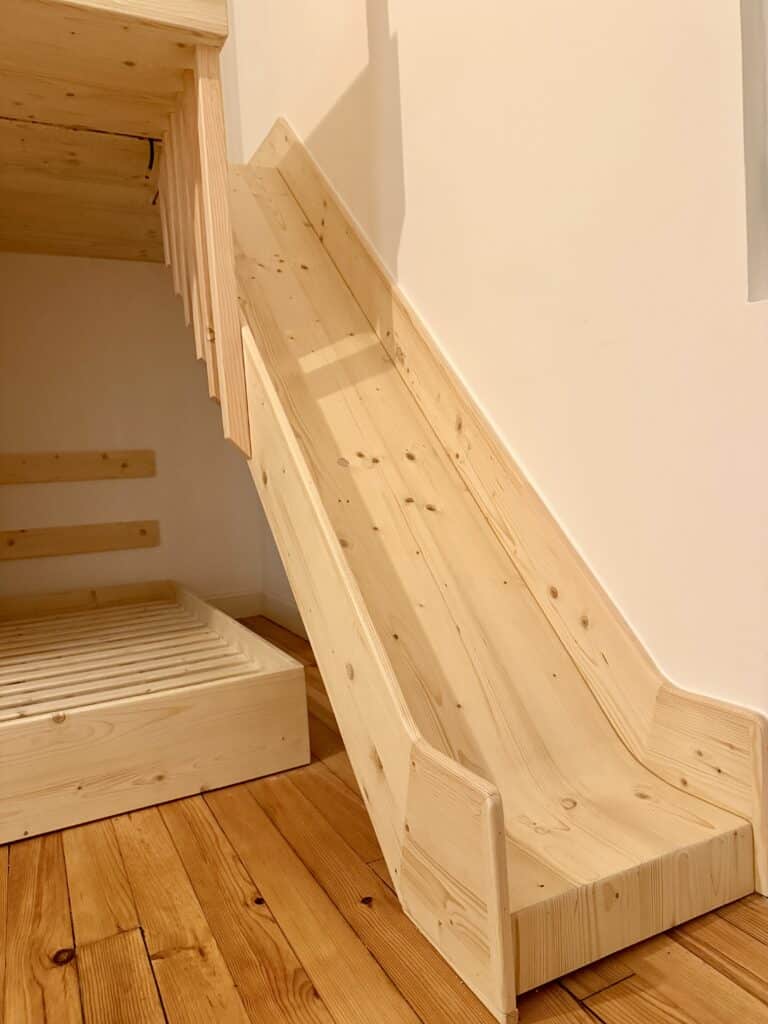Toboggan large en bois de pin neuf fixé à un lit superposé dans une chambre aux murs clairs. Plancher rustique.