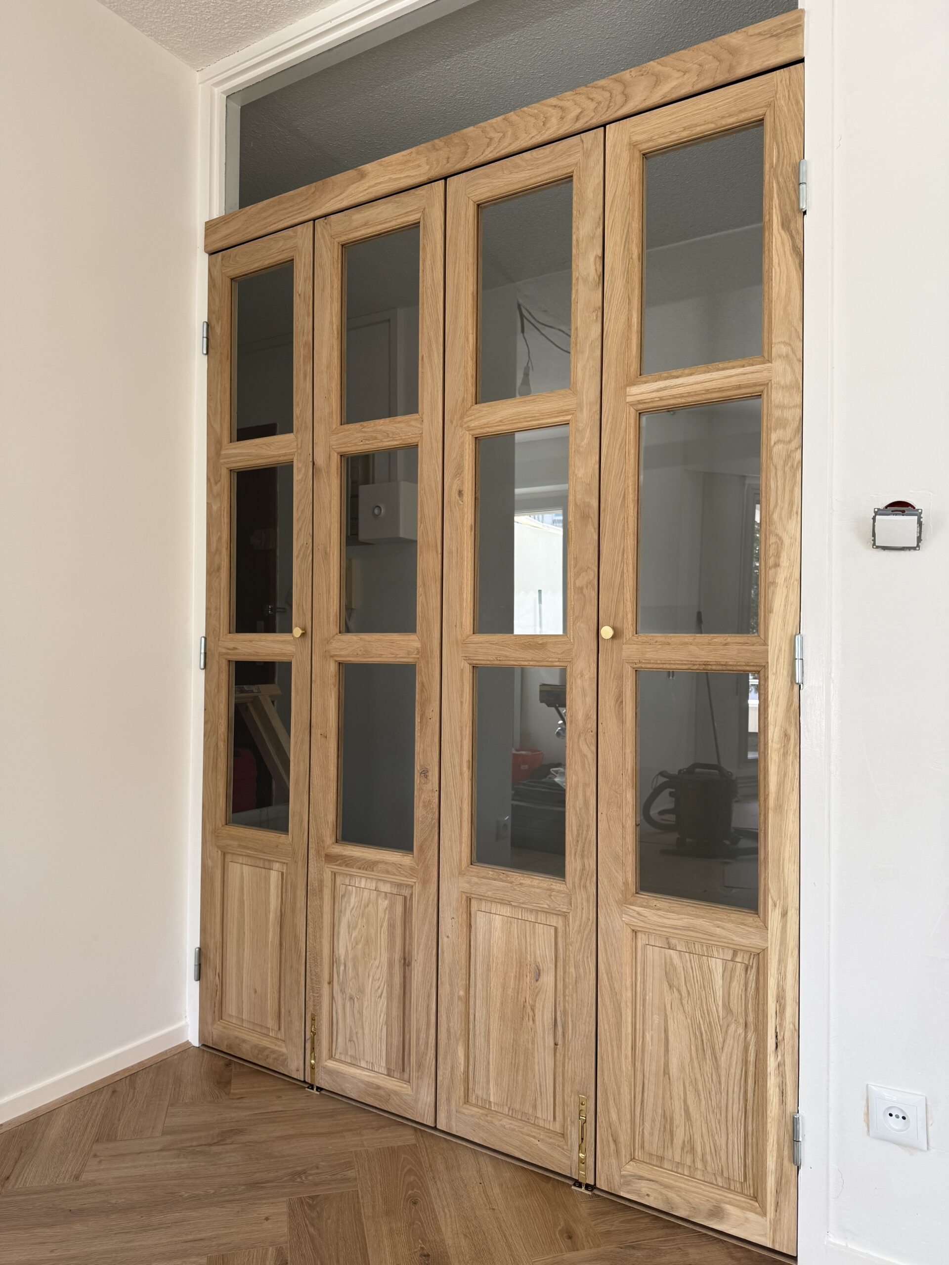 Porte accordéon vitrée en chêne massif style verrière installée sur un sol en parquet à chevrons.